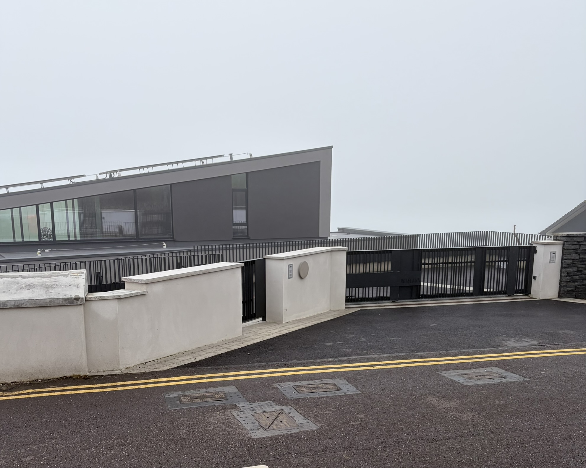 A modern glassy gray house behind gates on a foggy coastal road in Ballycotton
